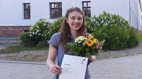 Die Studentin steh mit der Urkunde und einem Blumenstrauß in der Hand auf dem Campus.