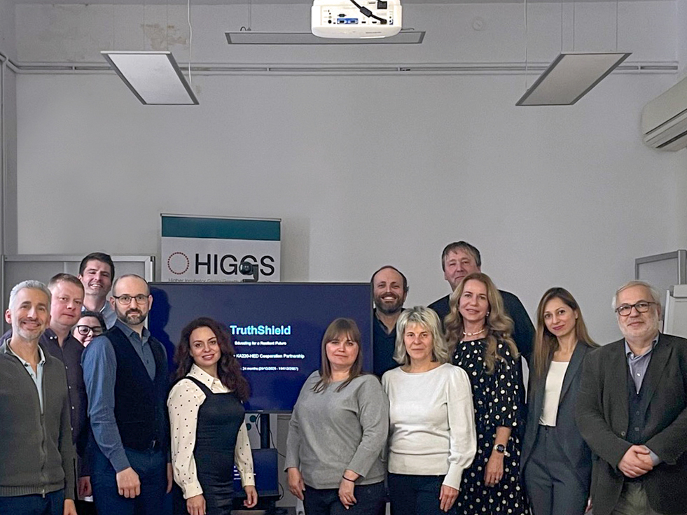13 Personen stehen nebeneinander in einem Seminarraum. Auf einem Bildschirm im Hintergrund steht "TruthShield".