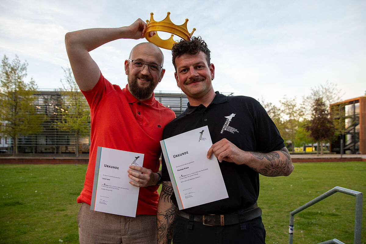 Die beiden stehen nebeinander. Sie halten in der linken Hand ihre Urkunden. Luca hält eine goldene Krone aus Plastik über beiden Köpfen.