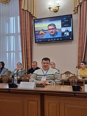 In-person participants seated in a conference room in Kyiv, with Prof. Dr. Arnaout joining virtually via screen in the background.