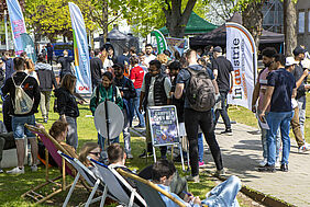 Viele junge Menschen laufen auf der Campuswiese zwischen Beachflages von ausstellenden Firmen. Im Vordergrund sitzen vier Personen in Liegestühlen. 