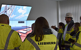 Man schaut zwei Teilnehmenden über die Schulter auf drei große Bildschirme, Darauf ist das Hafengelände zu sheen. Rechts steht der Mann, der erläutert, was zu sehen ist bwz. wie die Simulation abläuft.