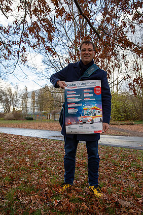 Professor Hillmann steht auf einer Wiese, die mit bunten Blättern zugedeckt ist. In den Händern hält er ein Plakat. Im Hintergrund sind Gebäude der Hochschule zu sehen.