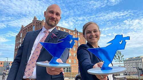 Stolz präsentieren Barbara Kienle und Florian Daniel die Innovationspreispokale, blaue Fischerfiguren, vor strahlend blauem Himmel mit herrlichem Wolkenmuster.