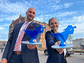 Stolz präsentieren Barbara Kienle und Florian Daniel die Innovationspreispokale, blaue Fischerfiguren, vor strahlend blauem Himmel mit herrlichem Wolkenmuster.