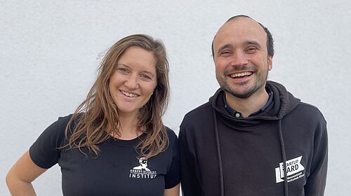 Carolin und Enrico stehen nebeneinander. Beide haben schwarze T-Shirts an. Auf Carolins steht "Robert Schmidt Institut" und auf Enricos "StartUpYard".