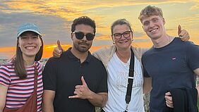 Vier Personen stehen vor einem Sonnenuntergang und lächeln in die Kamera, zwei Männer und zwei Frauen, eine trägt eine Kappe, eine Brille und eine Umhängetasche.