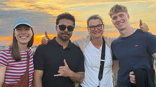 Vier Personen stehen vor einem Sonnenuntergang und lächeln in die Kamera, zwei Männer und zwei Frauen, eine trägt eine Kappe, eine Brille und eine Umhängetasche.