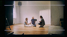 Alle drei Personen knien auf dem Boden in einem leeren Raum.