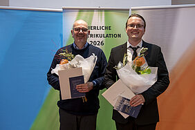 Zwei Männer in Anzügen stehen nebeneinander, halten jeweils einen Blumenstrauß und eine Urkunde, im Hintergrund ein Banner mit dem Text 'FEIERLICHE MATRIKULATION 2026'