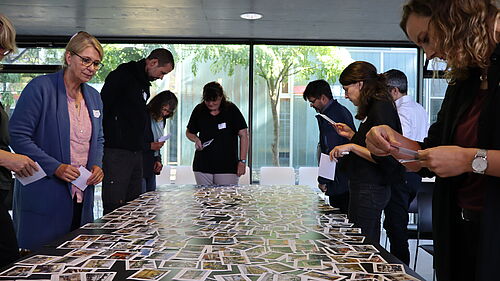 9 Personen stehen um einen Tisch erum und halten zum Teil Kartenin den Händen. Auf dem Tisch liegen Fotos im Postkartenformat.