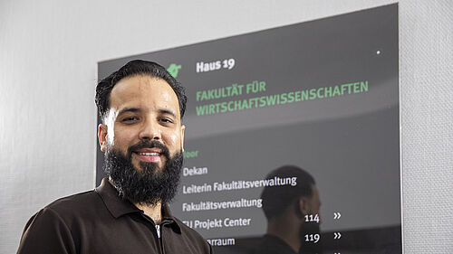 Mann mit Bart und dunklem Poloshirt steht vor schwarzem Bildschirm mit grüner und weißer Schrift im Fakultätsgebäude auf dem Campus