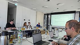 Es ist ein Raum zu sehen, in dem sich U-förmig angeordnete Tische befinden Daran sitzen fünf Personen, die sich Notizen machen - auf Papier und Laptops.
