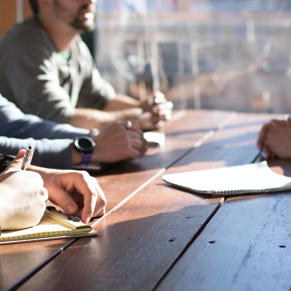 Nahaufnahme eines Holztisches, verschiedene Personen beugen sich über Dokumente und scheinen in eine Besprechung vertieft zu sein