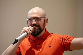 Es ist der Oberkörper des Mannes zu sehen. Mit der rechten Hand hält er ein Mikrofon an seiunen Mund, mit der linken zeigt er aus dem Bild heraus.