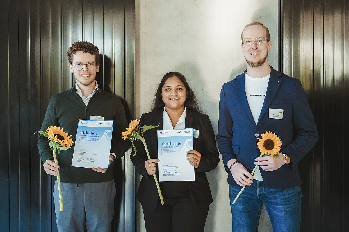 Die drei Personen stehen nebeneinander und halten je eine Urkunde und je eine Sonnenblume in den Händen.