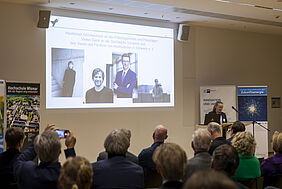 Über die Köpfe der TEilnehmenden hinweg schauen wir auf eine PRäsentation an der Wand. Darauf sind die Porträts der vier PResiträger zu sehen. Rechts am Bildrand ist die Laudatorin zu erkennen.