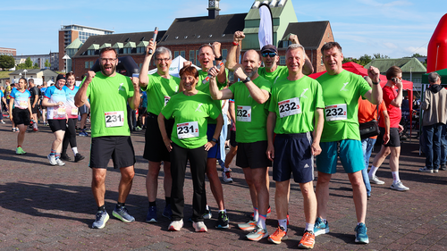 Eight participants from Wismar University of Applied Sciences, wearing green running shirts, pose together smiling and cheering before the start of the 16th Rostock Company Run. They wear team bibs numbered 231 and 232. Other runners, tents and the Haedge Peninsula architecture can be seen in the background.