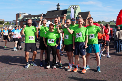 Acht Teilnehmende der Hochschule Wismar stehen in grünen Laufshirts beim Rostocker Firmenlauf 2025 zusammen, posieren gut gelaunt und mit erhobenen Armen für ein Gruppenfoto auf dem Veranstaltungsplatz. Sie tragen Startnummern mit den Teamnummern 231 und 232. Im Hintergrund weitere Läuferinnen und Läufer, Zelte und die Dächer der Haedgehalbinsel in Rostock.