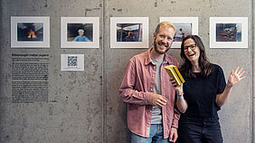 Die Beiden stehen vor einer Sichtbetonwand, an der sechs Fotos im fast A4-Format  zu sehen sind.