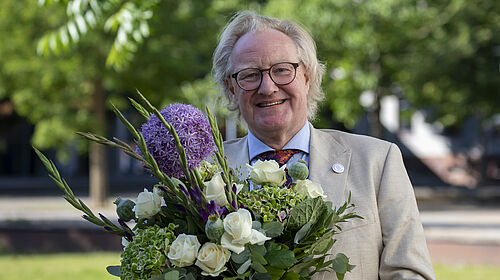 Der Rektor ist im Porträt auf dem Campus zu sehen. Er hält einen Blumenstrauß in der Hand.