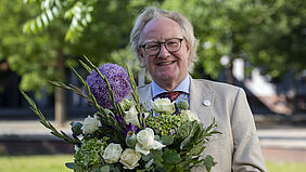 Der Rektor ist im Porträt auf dem Campus zu sehen. Er hält einen Blumenstrauß in der Hand.