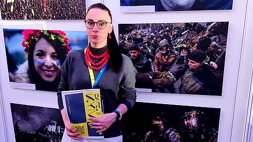 Dr Anna Fomenko, wearing a red necklace and holding a programme of the Cafe Kyiv event, stands in front of a wall of pictures reflecting impressions of the new reality of life for Ukrainian citizens. Between hope and suffering.