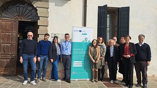 A group of project partners stands in front of a historic building next to a roll-up banner for the Interreg project ECLECTIC-CE and looks into the camera.