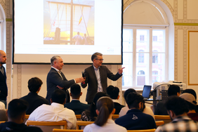 Two men stand in front of a seated group of students. One holds a microphone, the other is speaking. A presentation showing a sailing ship is displayed in the background.