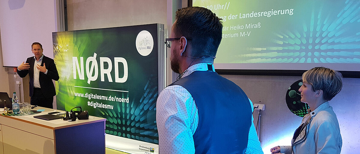 NØRD – Der erste landesweite Digitalisierungskrongress Im Vordergrund des Bildes stehen zwei Mitarbeiter des Innovation Ports der Hochschule Wismar und hören Heiko Miraß von der Landesregierung M-Vs bei seiner Rede auf dem landesweiten Digitalisierungskongress zu.