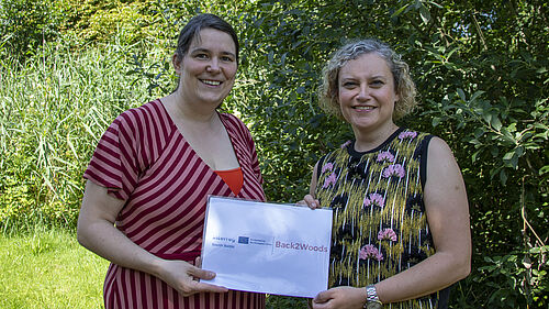 Die beiden Frauen stehen nebeneinander. Sie halten ein Schild in der Hand mit dem Titel des Projektes und den Logos der Fördermittelgeber. Im Hintergrund ist Wald zu sehen.