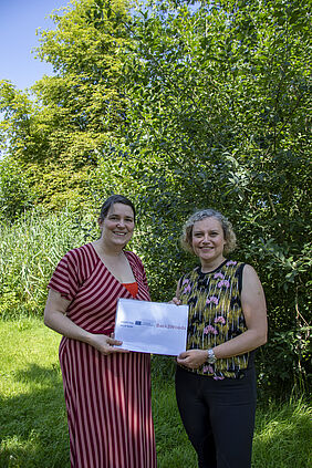 Die beiden Frauen stehen nebeneinander. Sie halten ein Schild in der Hand mit dem Titel des Projektes und den Logos der Fördermittelgeber. Im Hintergrund ist Wald zu sehen.