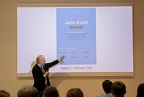 Der Moderator steht vor einer mit einem Beamer auf die Wand projezeirten Präsentation. Darauf ist ein Schild zu sehen.