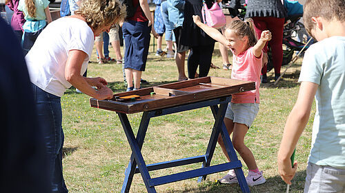 Ein Mädchen reißt vor Freude die Armen hoch. Sie steht an einem Kippeltisch mit einem Shuffelboard-ähnlichen Spiel. Die Oma ihr gegenüber schmunzelt.