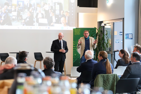 Zwei Männer stehen vor einem Publikum in einem modernen Veranstaltungsraum. Einer trägt einen schwarzen Anzug und hält ein Glas, der andere einen hellen Blazer und ein blaues Halstuch und spricht gestikulierend. Im Hintergrund ist eine Leinwand mit einer Videokonferenz sichtbar.