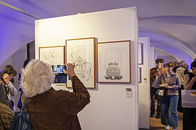Eine Person ist von hinten zu sehen, wie sie mit ihrem Handy die drei Grafiken von Louisa fotografiert. Die Grafiken hängen an einer Ausstellungstafel in einem weiß gestrichenen Festungsgewölbe. Dort sind noch weitere Besucher zu sehen.