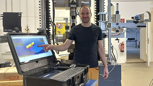 Mathias steht für einer Prüfmaschine, die viel höher als er ist. Er zeigt auf den Bildschirm eines Laptops. Darauf ist ein farbiges 3D-Diagramm zu sehen.