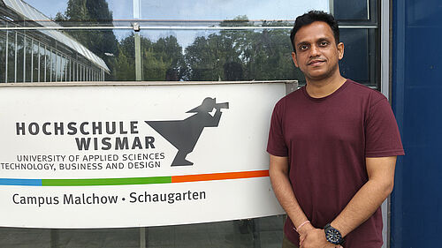Ein Mann steht vor einem Schild der Hochschule Wismar am Campus Malchow, das den Schriftzug „HOCHSCHULE WISMAR – University of Applied Sciences: Technology, Business and Design“ sowie das Logo der Hochschule zeigt. Im Hintergrund sind Glasfassaden und Bäume zu erkennen.