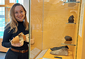 Es ist die Dozentin zu sehen, wie sie im Museum vor einer Vitrine steht und einen Holzschuh in der Hand hält.