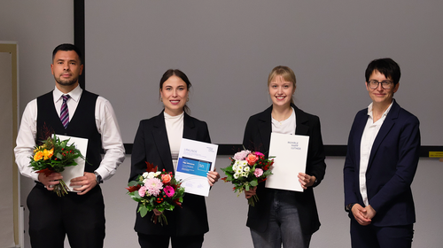 A woman in a dark blazer stands next to three young adults. All four are smiling and holding flower bouquets and certificates. The visible logos on the documents include Wismar University of Applied Sciences and Möhrle Happ Luther GmbH. The background shows a white wall with a projection screen.