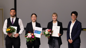 A woman in a dark blazer stands next to three young adults. All four are smiling and holding flower bouquets and certificates. The visible logos on the documents include Wismar University of Applied Sciences and Möhrle Happ Luther GmbH. The background shows a white wall with a projection screen.