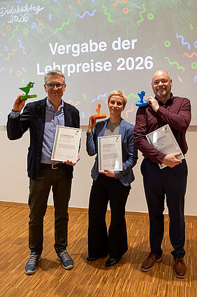 Die drei Personen stehen nebeneinander. Sie halten in der jeweils linken Hand eine Urkunde und  in Kopfhöhe einen kleinen Preispokal. Dieser hat jeweils die Farbe der Fakultät: Grün, Orange und Blau.