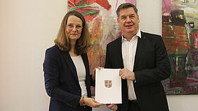 Die Ministerin und der Professor halten die Ernennungsurkunde in der Hand. An der Wand hinter ihnen hängen Bilder.