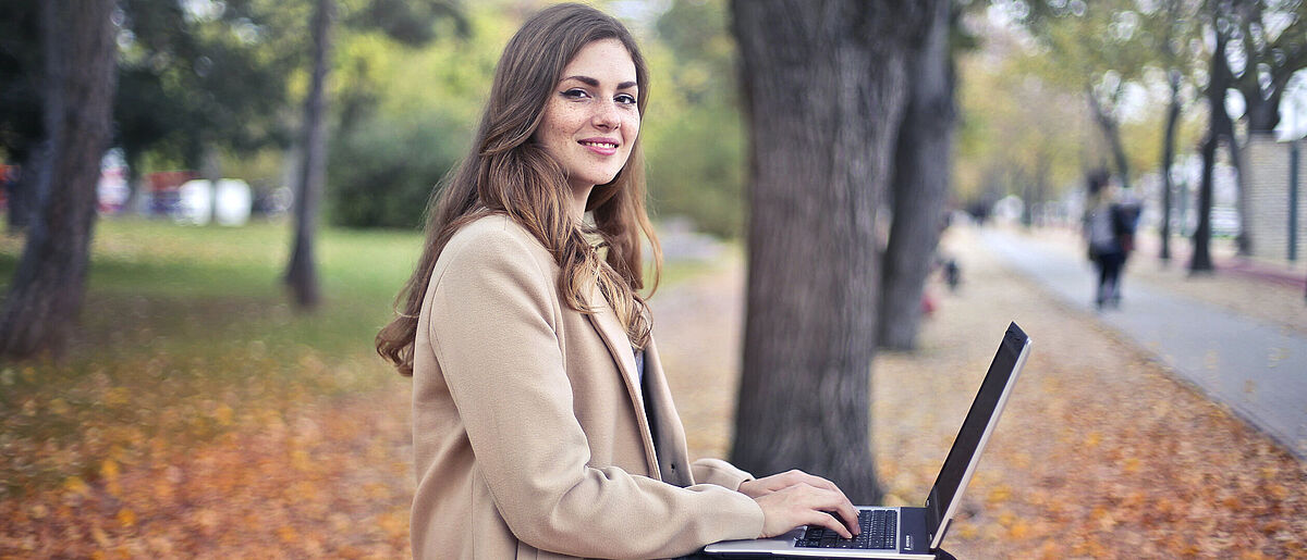 Jetzt bewerben! Junge Frau im Park mit Laptop auf dem Schoß lächelt in die Kamera