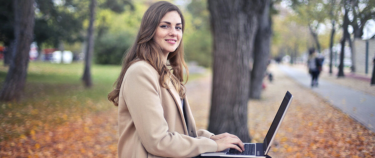 Jetzt bewerben! Junge Frau im Park mit Laptop auf dem Schoß lächelt in die Kamera