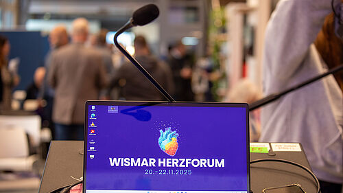 Auf dem Bildschirm eines Laptops ist eine Herzgrafik zu sehen. Dahinter im Raum sind unscharf Menschen im Gespräch zu erkennen.