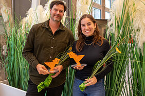 Zwei lächelnde Personen halten orangefarbene Fischerpokale und Blumensträuße vor hohen grünen Pflanzen.