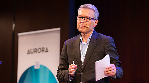 A speaker stands on stage wearing a headset microphone and gives a presentation in front of an Aurora banner at the Aurora Symposium on Global Citizenship 2026.