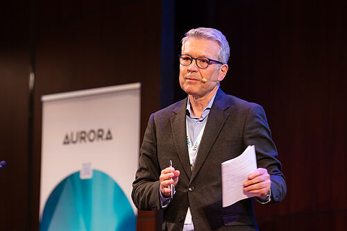 A speaker stands on stage wearing a headset microphone and gives a presentation in front of an Aurora banner at the Aurora Symposium on Global Citizenship 2026.