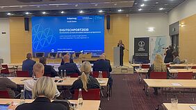 A woman speaks at a lectern in front of a large screen showing the DigiTechPort2030 presentation. Conference attendees sit at tables in the foreground.